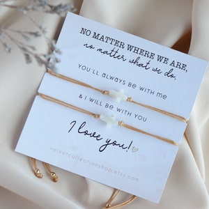 May include: Two beige string bracelets with white cross charms. The bracelets are tied together with a card that reads "NO MATTER WHERE WE ARE, no matter what we do, YOU'LL ALWAYS BE WITH ME & I WILL BE WITH YOU I love you!"