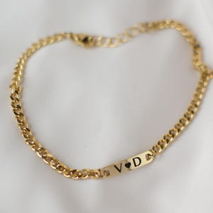 May include: Gold-colored chain bracelet with a rectangular plate. The plate features the engraved letters "V D" with a heart symbol. The bracelet is displayed on a white, textured surface.