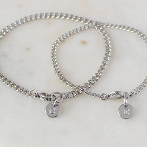 May include: Two silver-colored chain bracelets with small, round charms. One charm has the letter "C" and the other has the letter "E". The bracelets are displayed on a white marble surface.