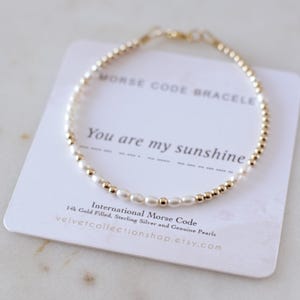 以下が含まれることがあります： 白いカードに「You are my sunshine」というメッセージが書かれた、金色と白のビーズブレスレット。ブレスレットは14金メッキのスターリングシルバーと本物の真珠で作られています。カードには「International Morse Code」と「velvetcollectionshop.etsy.com」の文字も書かれています。