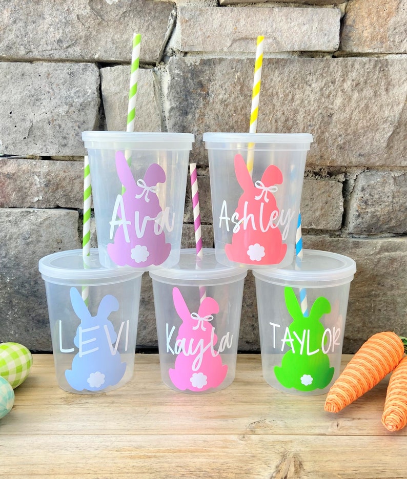 May include: Five clear plastic cups with straws and bunny designs. The cups have the names Ava, Ashley, Levi, Kayla, and Taylor written on them. The bunnies are pink, blue, green, and pink. The cups are sitting on a wooden surface with carrots and Easter eggs in the background.