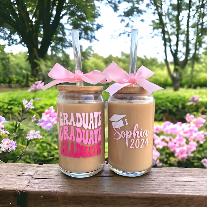 May include: Two glass cups with lids and straws. The cups are decorated with pink and white designs. One cup has the text 'Graduate' repeated three times in a pink and white gradient. The other cup has the text 'Sophia 2024' and a graduation cap design.