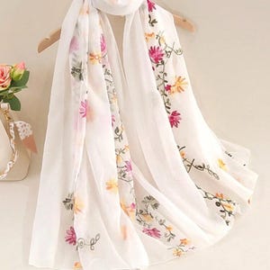 Puede incluir: Bufanda blanca con bordados florales en rosa, amarillo y verde. La bufanda está drapeada sobre una percha de madera, mostrando su tejido ligero y fluido. El diseño floral añade un toque de elegancia.