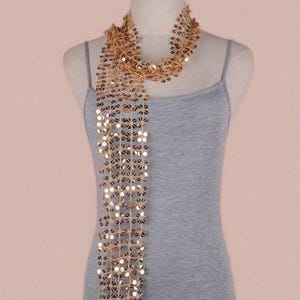 May include: A gold sequin scarf draped over a grey vest top on a mannequin. The scarf is made of small, round, gold sequins and hangs down in vertical strands. The vest top is a simple, sleeveless design.