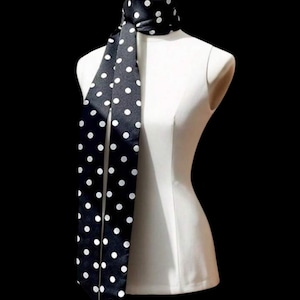 Puede incluir: Una bufanda negra con lunares blancos está drapeada sobre un maniquí blanco. La bufanda está hecha de un material brillante y tiene un patrón clásico de lunares. El fondo es negro.