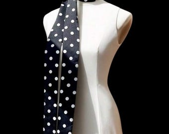 May include: A black scarf with white polka dots is draped over a white mannequin. The scarf is made of a shiny material and has a classic polka dot pattern. The background is black.