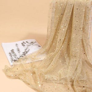 May include: A sheer gold fabric with small gold sequins scattered throughout. The fabric is draped over a book and a white surface.