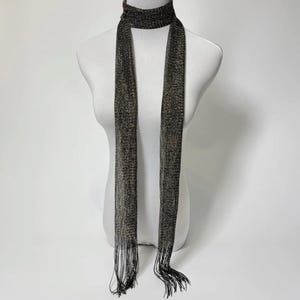 Puede incluir: Bufanda metálica estrecha con flecos en los extremos. La combinación de colores es negro y dorado. Se muestra sobre un maniquí para resaltar su caída elegante. Un accesorio de moda versátil para complementar cualquier atuendo.