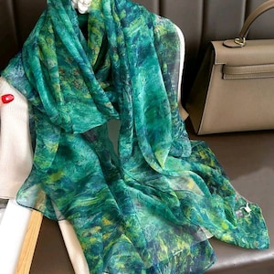May include: A sheer green scarf with a watercolour-like pattern of blues and greens. The scarf is draped over a white sweater and a wooden hanger. A tan handbag is visible in the background.