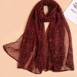 Può includere: Una sciarpa rossa trasparente con paillettes e una finitura scintillante. La sciarpa è piegata a metà e drappeggiata su una superficie marrone chiaro.