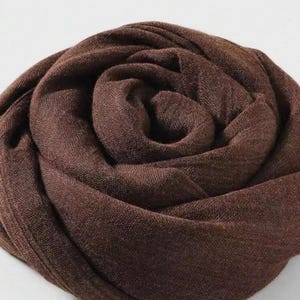 May include: A rolled, dark brown scarf. The fabric appears to be a soft, woven material. The scarf is arranged in a spiral, with the folds creating a textured, layered effect. The color is a rich, earthy tone.