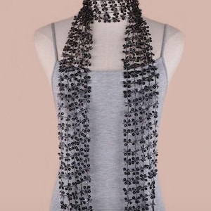 Puede incluir: Bufanda negra con cuentas con un cuello tipo gargantilla y hebras largas y fluidas. La bufanda se exhibe en un maniquí que lleva una camiseta sin mangas gris. Las cuentas son pequeñas y crean un delicado patrón floral.