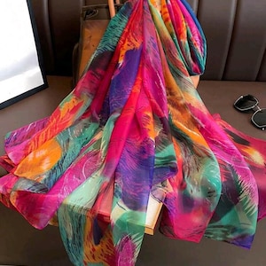 Puede incluir: Una bufanda vibrante y colorida con un diseño similar a plumas. La bufanda presenta una mezcla de colores brillantes, incluyendo rosa, morado, naranja y verde. La bufanda está drapeada sobre un bolso de cuero marrón, creando un accesorio elegante y llamativo.