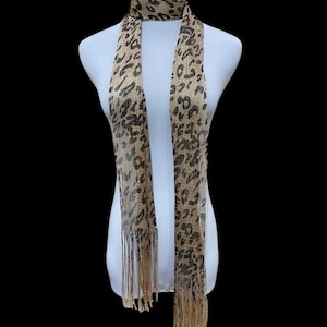 May include: A tan and black leopard print scarf with fringe. The scarf is draped over a white mannequin. The scarf has a repeating pattern of black spots on a tan background. The fringe is a light tan color.