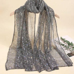 May include: A grey scarf with a sheer, shimmering fabric. The scarf is embellished with small, iridescent sequins. It is draped over a wooden hanger, highlighting its flowing design. A fashionable accessory, perfect for adding a touch of glamour.