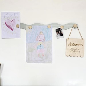 May include: A pale blue wooden display board with a wavy design, displaying children's drawings and a small photograph. The artwork includes a drawing of a heart with the word "Mum" and a drawing of a person. A wooden sign reads "Autumn's Creative Corner."