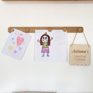 May include: A wooden display board with three drawings and a sign. The drawings include a heart, a princess, and a handwritten note. The sign reads "Autumn's Creative Corner". The board is made of light brown wood.
