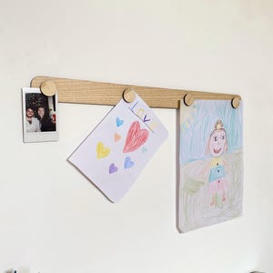 May include: A light-coloured wooden picture rail with three wooden pegs holding a photo and two children's drawings. The drawings include a heart with the word "LOVE" and a drawing of a person with a crown. Coloured pencils are visible at the bottom.