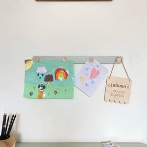 May include: A children's art display with a green and grey wooden picture rail holding artwork and a wooden sign reading "Autumn's Creative Corner." A sign above reads "Reading Corner."