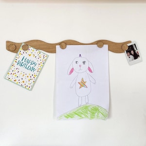 May include: A wooden picture holder with a wavy design, holding a birthday card, a child's drawing of a unicorn, and a small photograph. The birthday card has the text "HAPPY BIRTHDAY" with colorful confetti.