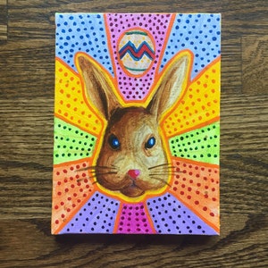 Peut inclure: Une peinture colorée d'un lapin brun sur un fond rose, bleu, jaune, vert et orange. Le lapin a un nez rose et des moustaches blanches. Il y a un œuf de Pâques rose avec un motif bleu et rouge sur la tête du lapin.