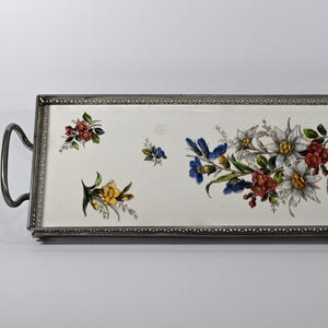 Puede incluir: Bandeja rectangular con superficie de cerámica blanca y marco de metal plateado. La bandeja está decorada con coloridos estampados florales, incluyendo flores azules, rojas y amarillas. Tiene asas curvas en cada extremo.