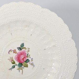 Könnte beinhalten: Weißer Keramikteller mit einem erhabenen Spitzenmuster am Rand. Die Mitte des Tellers zeigt eine handgemalte rosa Rose mit grünen Blättern und anderen Blumen.