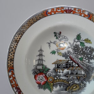 May include: A white ceramic plate with a black, red, and gold border. The center of the plate features a black and white illustration of a pagoda, a tree, and a house with a red flower.