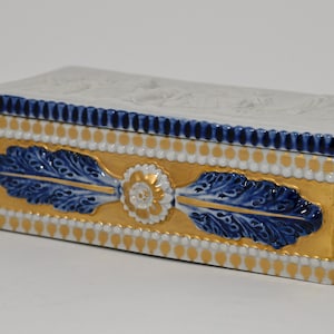 Könnte beinhalten: Rechteckige Porzellandose mit weißem Deckel mit geprägten Details. Die Dose hat einen goldenen Sockel mit blau-weißen floralen Designs und einem blauen Rand. Die Dose ist mit goldenen Akzenten verziert.