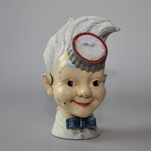 Può includere: Una statuetta in ceramica bianca di un ragazzo con un tappo di bottiglia bianco e rosso sulla testa. Il ragazzo indossa una camicia bianca con un papillon blu.