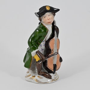 May include: A porcelain figurine of a monkey dressed in a green coat and black hat, playing a cello. The monkey has a brown face and is standing on a white base.