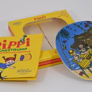 K&ouml;nnte beinhalten: Eine gelbe Schachtel und ein Teller mit einer Cartoon-Illustration von Kindern am Strand. Der Teller ist blau mit einer Strandszene. Ein gelbes Buch mit dem Titel "Pippi L&aring;ngstrump" befindet sich ebenfalls im Bild.