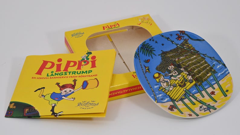 K&ouml;nnte beinhalten: Eine gelbe Schachtel und ein Teller mit einer Cartoon-Illustration von Kindern am Strand. Der Teller ist blau mit einer Strandszene. Ein gelbes Buch mit dem Titel "Pippi L&aring;ngstrump" befindet sich ebenfalls im Bild.