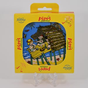 K&ouml;nnte beinhalten: Eine gelbe quadratische Schachtel mit einer farbenfrohen Illustration von Pippi Langstrumpf und ihren Freunden am Strand. Auf der Schachtel stehen die Worte "Pippi L&aring;ngstrump" und "R&ouml;rstrand Sweden". Das Bild zeigt eine Strandszene mit einer H&uuml;tte und Kindern.
