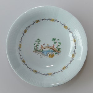 May include: A white ceramic plate with a blue and yellow border design. The center of the plate features a hand-painted scene of a bridge with two figures in a landscape setting.