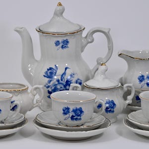 Puede incluir: Un juego de té de porcelana blanca con diseños florales azules. El juego incluye una tetera, azucarero, jarra de crema y varias tazas de té con platillos. Los bordes de cada pieza están adornados con detalles dorados, creando un aspecto elegante.