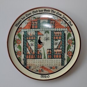 May include: A white ceramic plate with a colorful illustration of a building with people inside. The plate has a brown rim and the text "Nineve den Store Stad han Gord Bot Com Jonas Bod" and "MRD" are printed on the plate.
