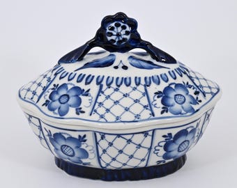 May include: A white and blue ceramic lidded serving dish. The dish has a floral design with blue accents and a lattice pattern. The lid has a dark blue handle with a flower design.