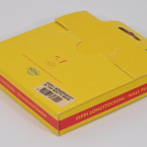 K&ouml;nnte beinhalten: Gelbe Pappschachtel mit rotem Rand, mit Text in mehreren Sprachen. Die Schachtel hat einen ausgeschnittenen Griff und ist mit "Pippi Longstocking Wall Plate" beschriftet. Die Schachtel ist geschlossen und angewinkelt.