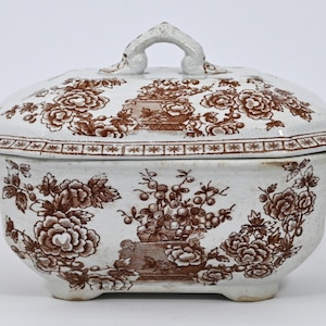 Puede incluir: Un plato de servir de porcelana blanca con tapa. El plato está decorado con un patrón floral marrón. La tapa tiene un asa en la parte superior.