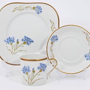 May include: A white porcelain dish set featuring a square plate, a round plate, and a mug. Each piece is adorned with blue floral designs and gold trim. The set is displayed on clear stands, showcasing the elegant design.