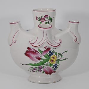 May include: A white ceramic tulip vase with a wide, rounded body and three spouts. The vase is decorated with pink and red floral designs, including tulips and other flowers. The edges are trimmed with a pink border.
