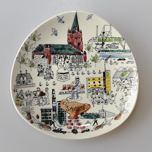 以下が含まれることがあります： 白いセラミック製のプレートに、カラフルな街の風景が描かれています。イラストには、教会、建物、人々、そして「Savdnes」という言葉が含まれています。