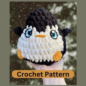 Può includere: Schema di uncinetto per un giocattolo amigurumi di pinguino. Il pinguino è bianco con accenti neri e ha grandi occhi blu. Il giocattolo è tenuto in una mano.