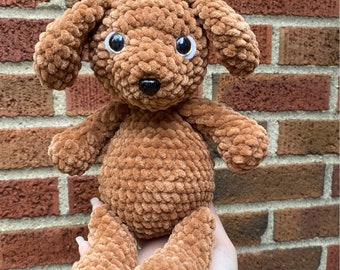 Puede incluir: Un juguete de peluche de perro de crochet marrón con grandes ojos negros y una nariz negra. El perro está sentado en una mano.
