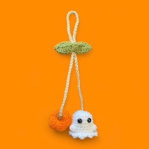 Könnte beinhalten: Handgefertigter Halloween-Häkelornament. Es zeigt einen weißen Geist mit schwarzen Augen und einen orangefarbenen Kürbis, der an einem grünen Blatt und einer weißen Kordel hängt. Der Hintergrund ist orange. Ideal für saisonale Dekorationen.