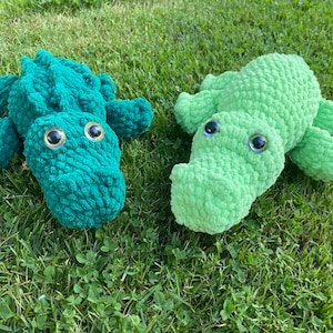 May include: Two crocheted alligator toys, one green and one light green, with black eyes, lying on a bed of green grass.