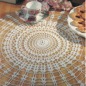 May include: White crocheted doily with a delicate, intricate design. The doily is laid on a wooden table with a cup and saucer and a plate of cookies.