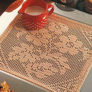 May include: A peach-colored, crocheted placemat with a geometric design. The placemat is square and has a detailed openwork pattern. A red pitcher with white heart-shaped designs is visible in the upper left corner, filled with a white liquid.