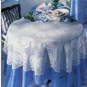 Puede incluir: Un mantel de crochet blanco con un diseño floral, colocado sobre un mantel azul con un patrón de conchas marinas. El mantel está sobre una mesa redonda con una base blanca. Hay dos vasos y un plato de comida en la mesa.